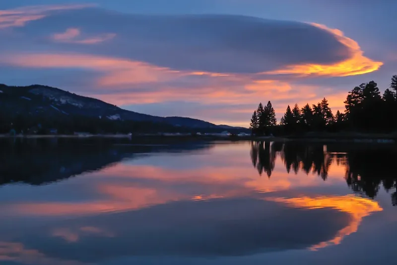 Pink and orange sunrise clouds and pines reflected in still water at Big Bear Lake