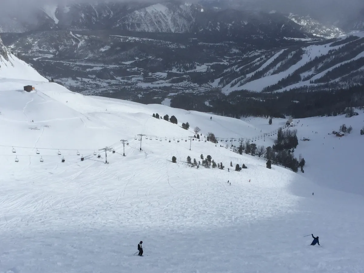 Two skiers on a wide, gentle snowy run at Big Sky Resort, with a chairlift line, scattered pines and a forested valley below