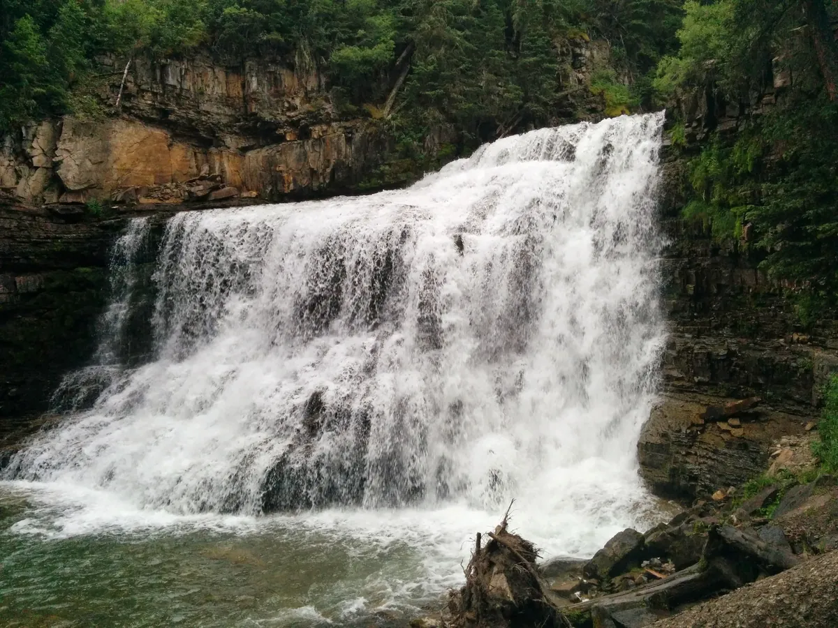 Ousel Falls at Big Sky in summer, a wide waterfall pouring over a rock ledge into a pool, with pines on the canyon walls