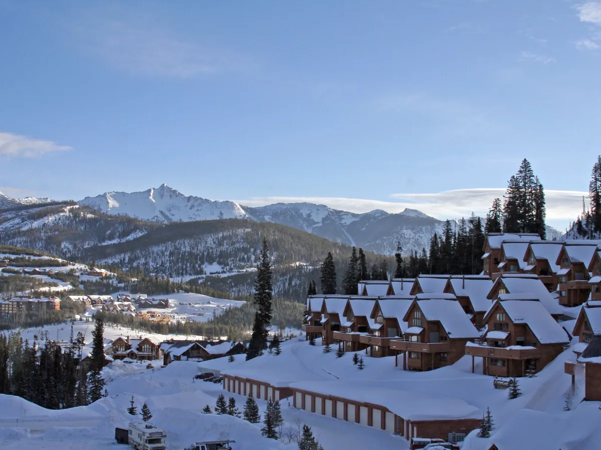 Snow-covered lodges and condos of Big Sky Resort’s Mountain Village among pines, with Lone Peak rising white behind under a blue sky