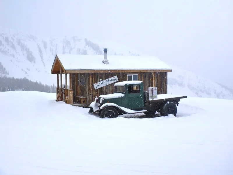 A snowy log cabin in Big Sky with an old pickup truck half-buried in deep snow beside it