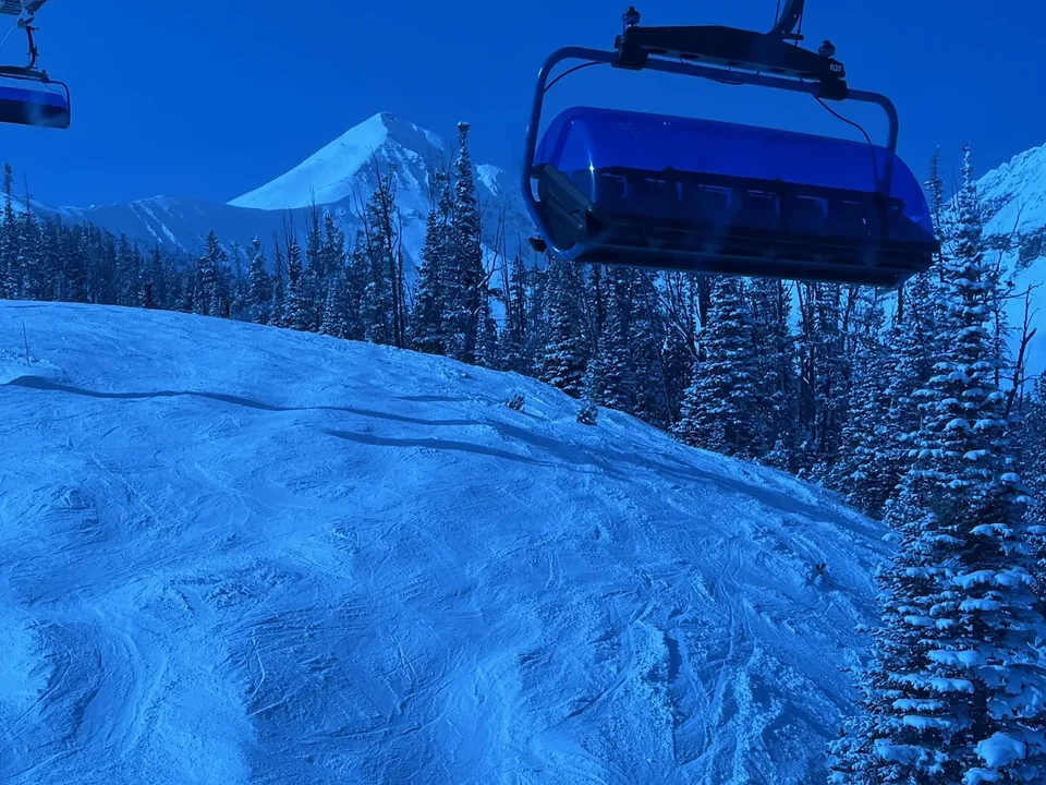 A chairlift with a blue weather bubble passing over a groomed run at Big Sky Resort, with snowy pines and a white peak behind