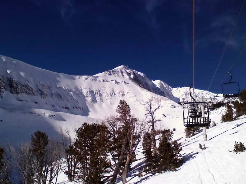 A view up a chairlift line toward the rocky summit of Lone Peak, with snowy slopes and trees below