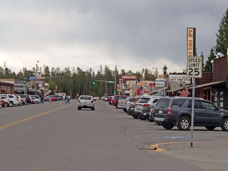 A street in downtown West Yellowstone with low shops, signs and parked cars under a cloudy sky