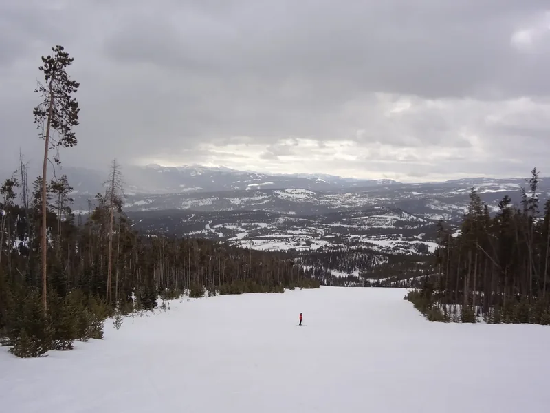 A wide snowy ski run at Big Sky seen from the top, with pines and a valley of ridges below under cloud