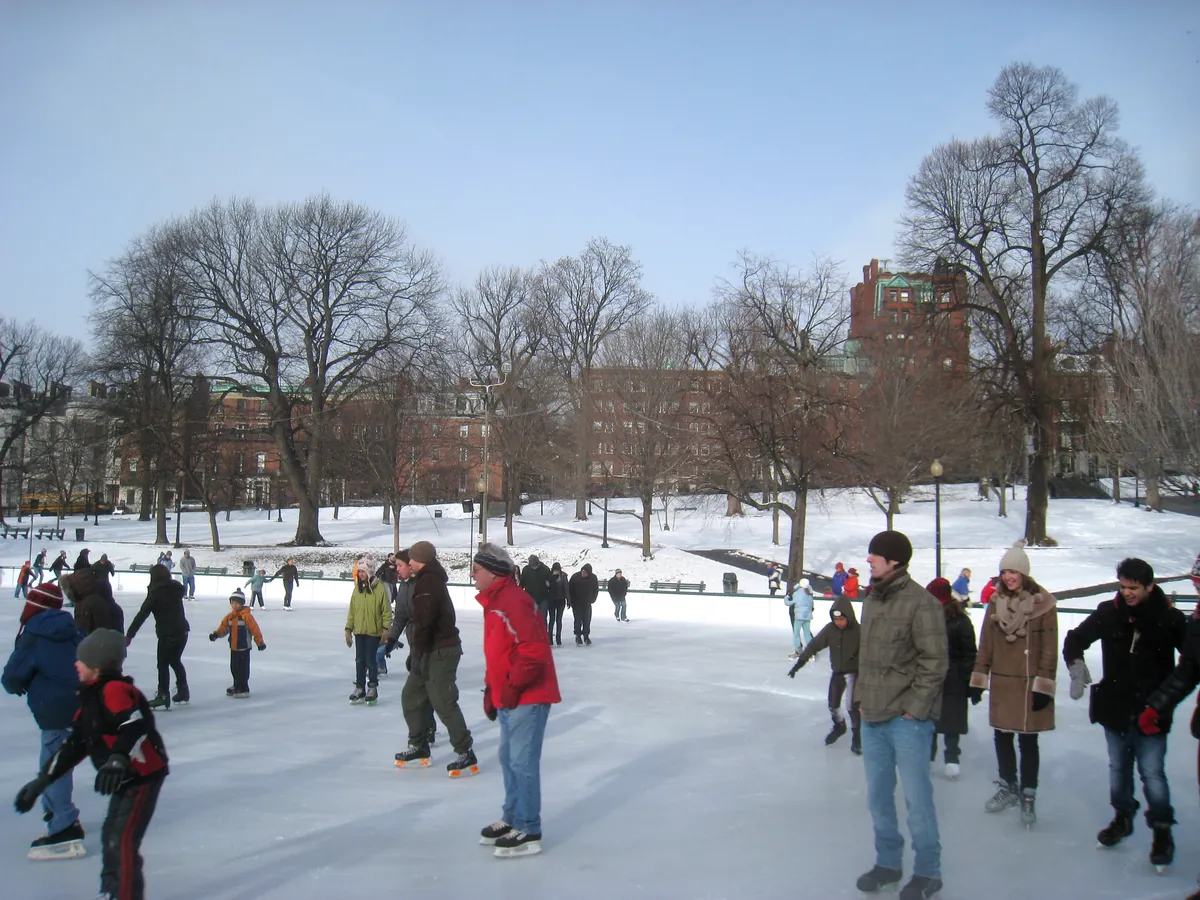 Skaters of all ages circling the Frog Pond rink on snowy Boston Common under bare trees, with brick buildings beyond