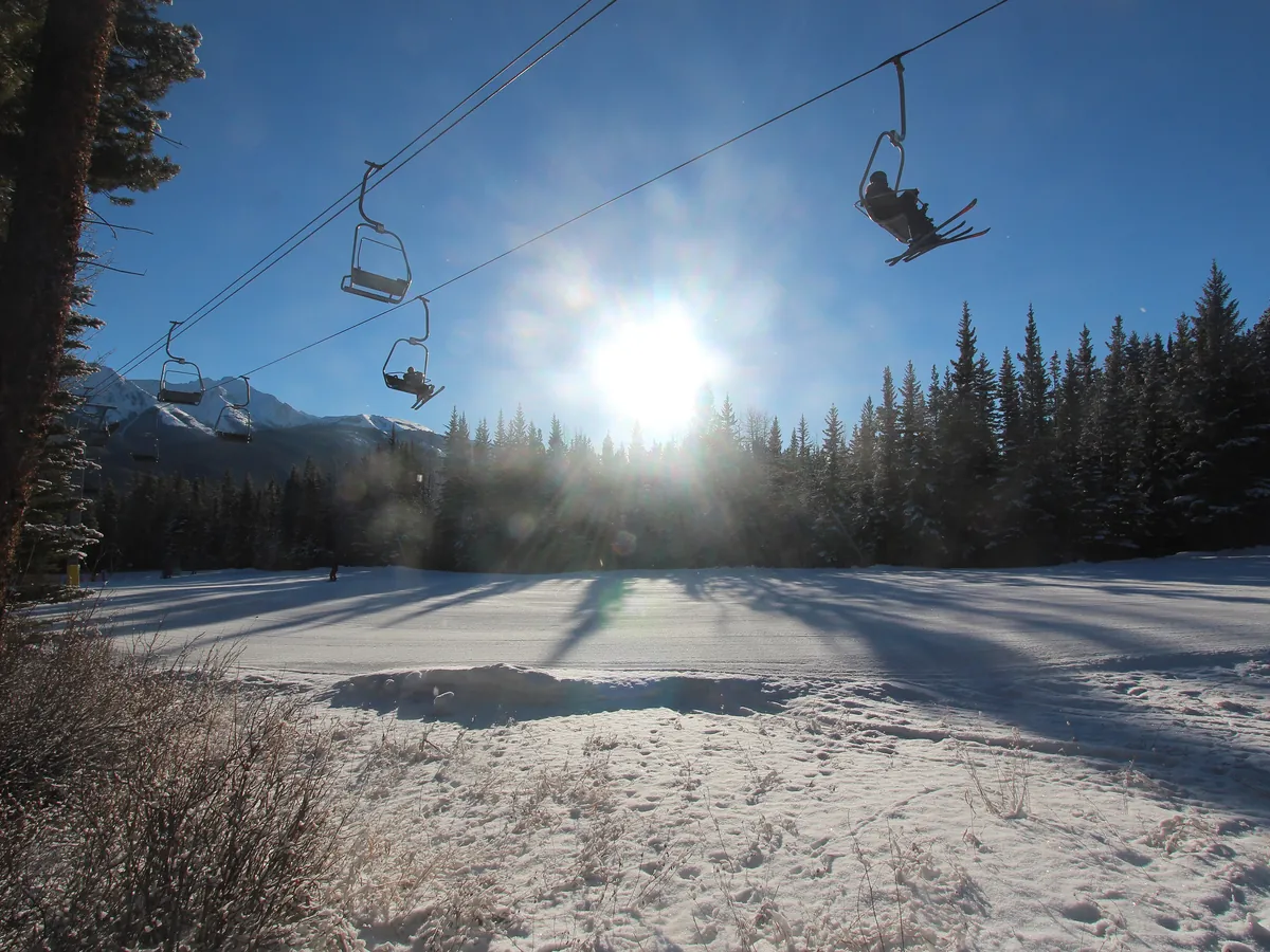 A chairlift at Nakiska rising over a snowy slope with the sun low behind the trees