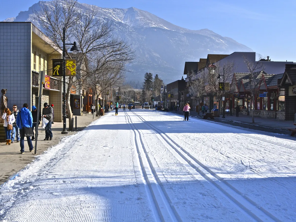 A snowy street in Canmore with ski tracks down the middle, low shops and people walking, under a snowy mountain