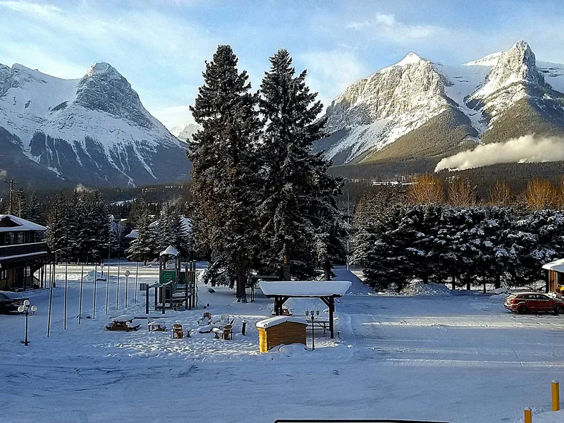 A snowy park and playground in Canmore with tall spruce trees and snow-covered peaks behind under morning sun