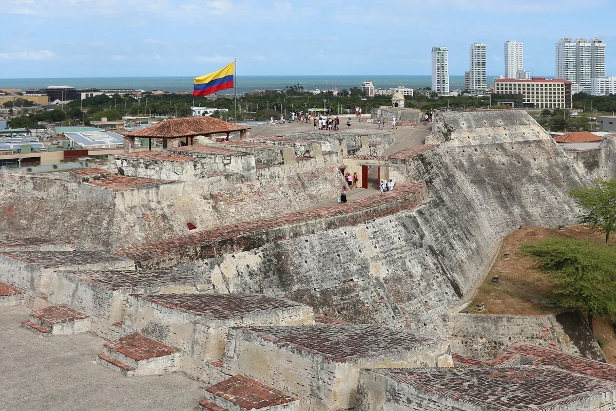 The stone ramparts of Castillo San Felipe de Barajas with a Colombian flag and city towers beyond