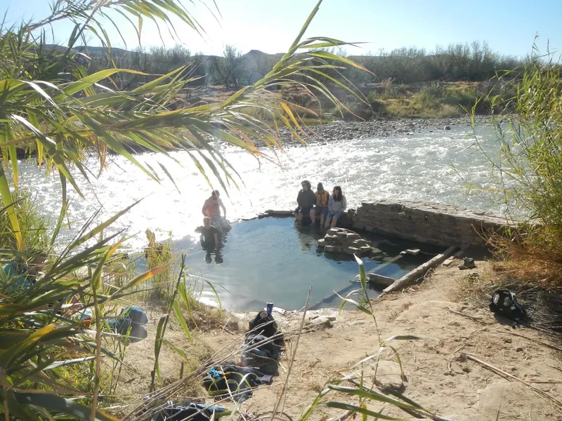 People sitting in the stone-walled hot springs pool beside the Rio Grande, with palm fronds in the foreground