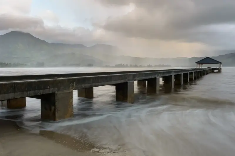 The long wooden Hanalei Pier reaching into Hanalei Bay under low clouds and green mountains