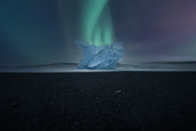 A glassy chunk of ice on black sand at Diamond Beach under green aurora