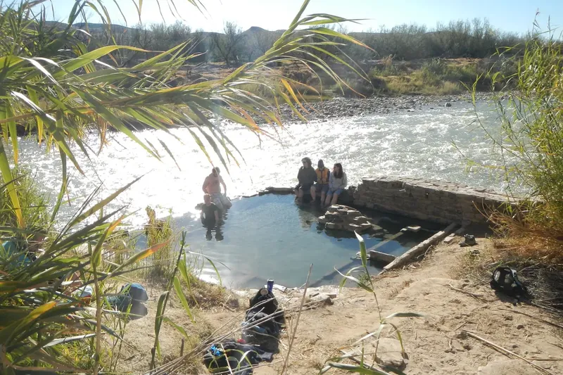 People sitting in the stone-walled hot springs pool beside the Rio Grande, with palm fronds in the foreground