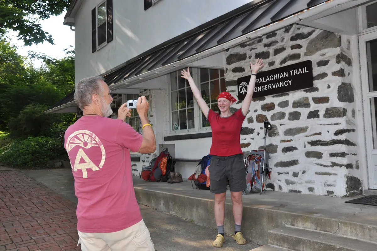 A hiker posing on the porch of the Appalachian Trail Conservancy building in Harpers Ferry while a staff member takes a photo