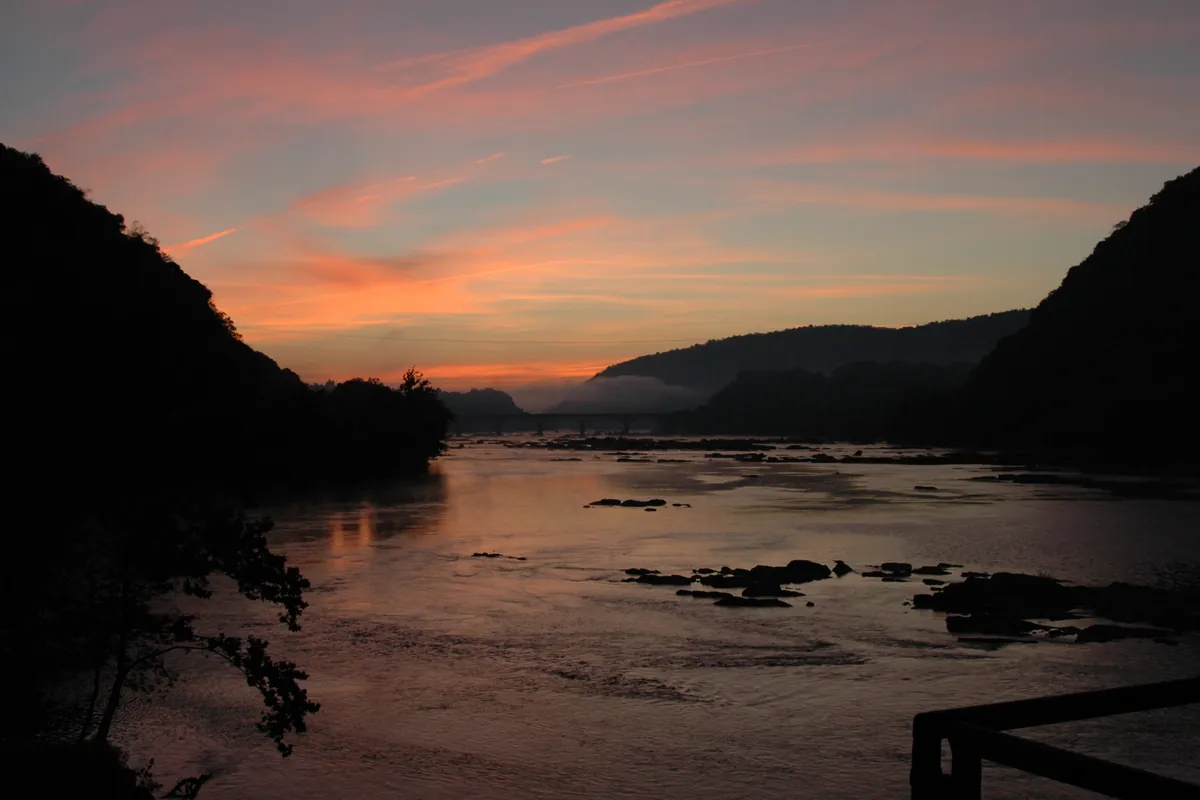 A river at dusk under an orange sky, with dark wooded ridges on both sides near Harpers Ferry