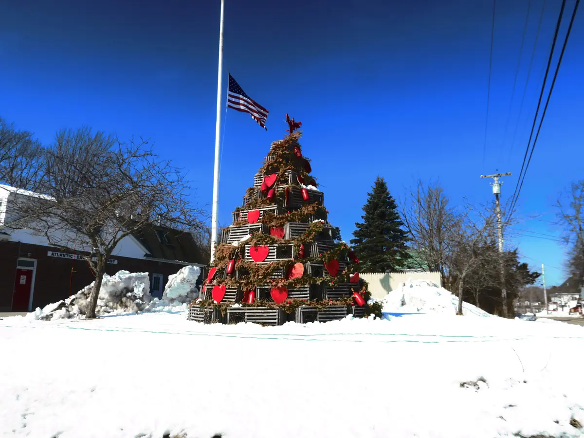 A Christmas tree built from stacked lobster traps and buoys in snowy Cape Porpoise Square, with a flagpole and blue sky