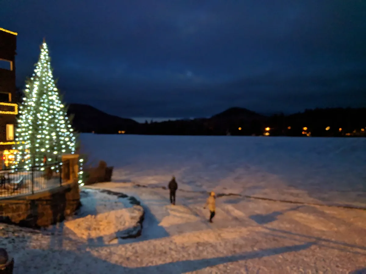Snow-covered frozen Mirror Lake at dusk, with a lit Christmas tree on the shore and the hills beyond