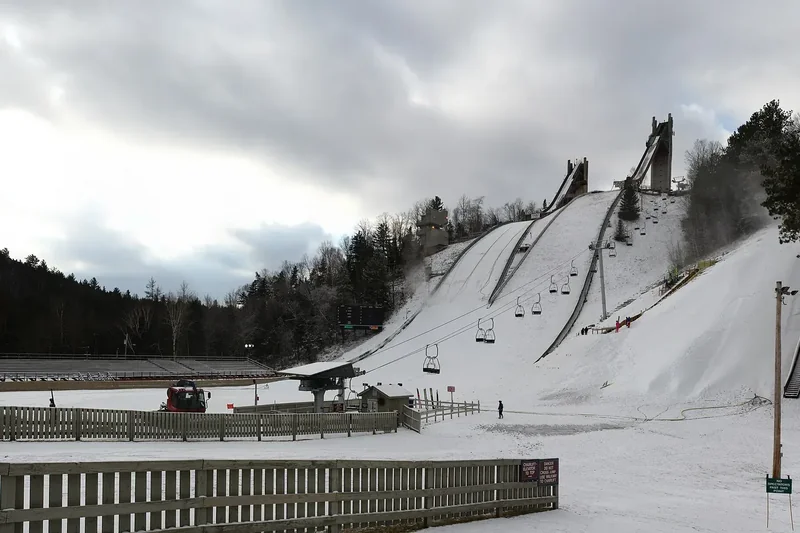 The Olympic ski jump towers at Lake Placid above a snowy landing hill, a chairlift and wooden fence in front