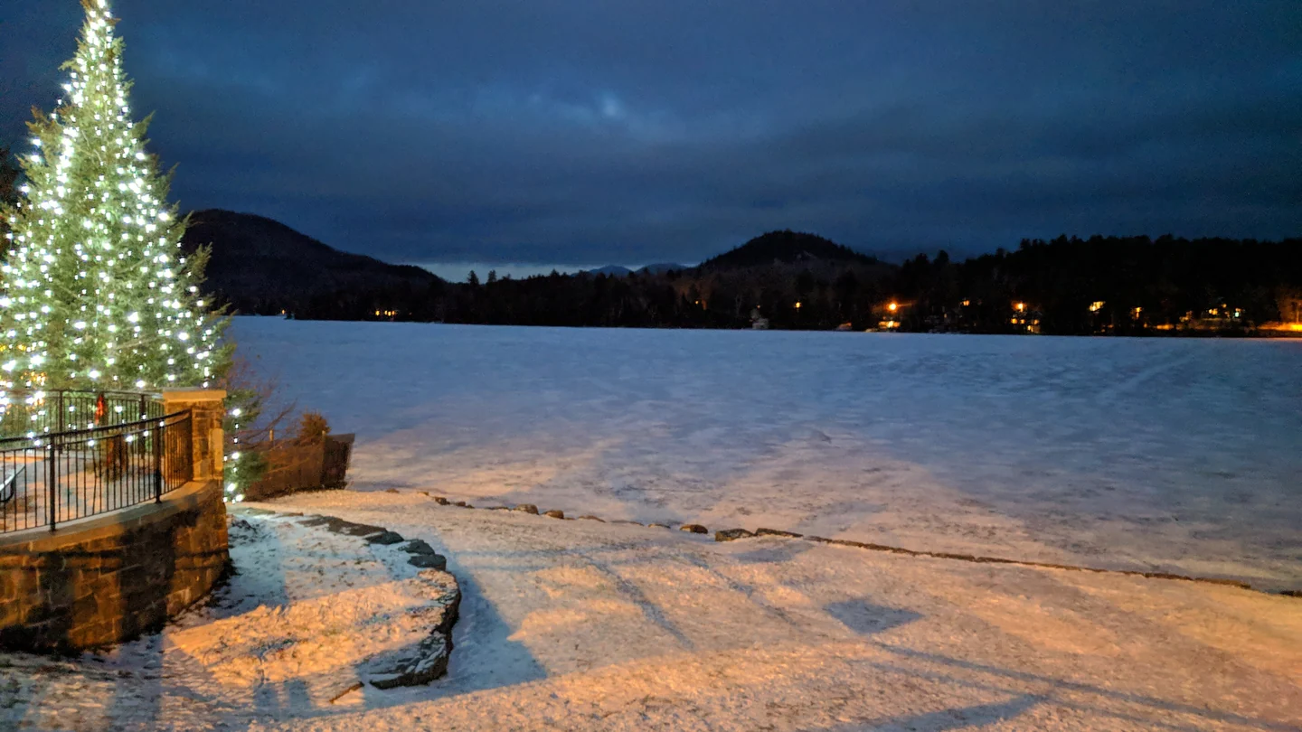 A lit Christmas tree beside snow-covered Mirror Lake in Lake Placid at dusk, hills beyond