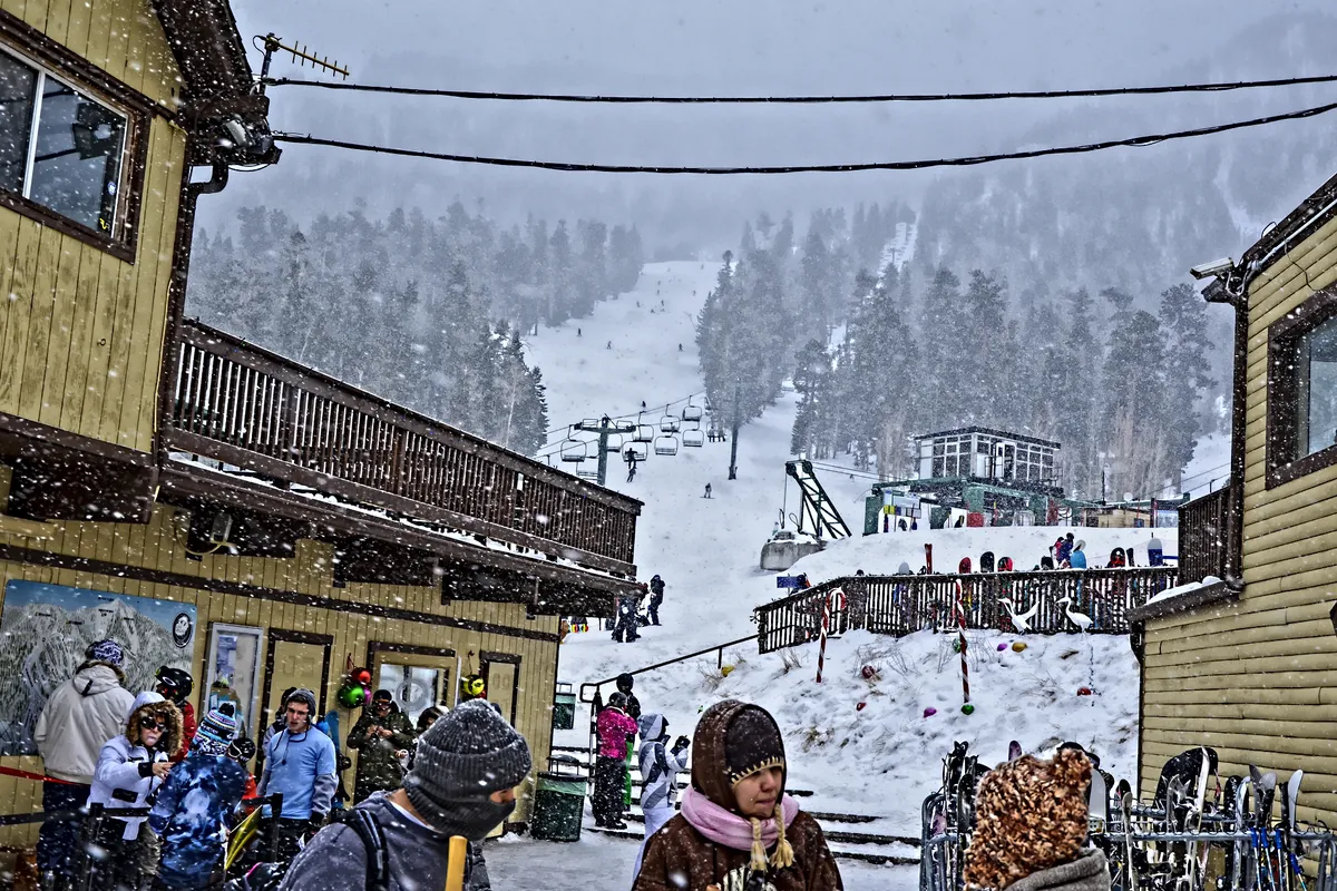 Visitors in winter coats on a snowy deck at the Lee Canyon base area, with ski runs and pines behind