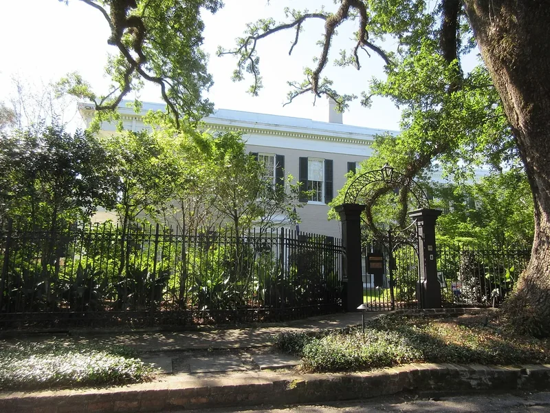 A white Garden District house behind an iron fence and green oaks in New Orleans