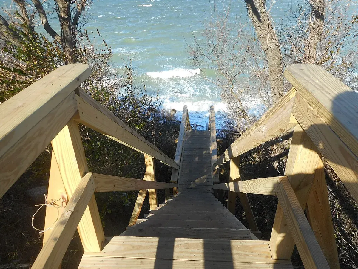 A wooden stair with handrails leading down a wooded bluff toward the water of Long Island Sound