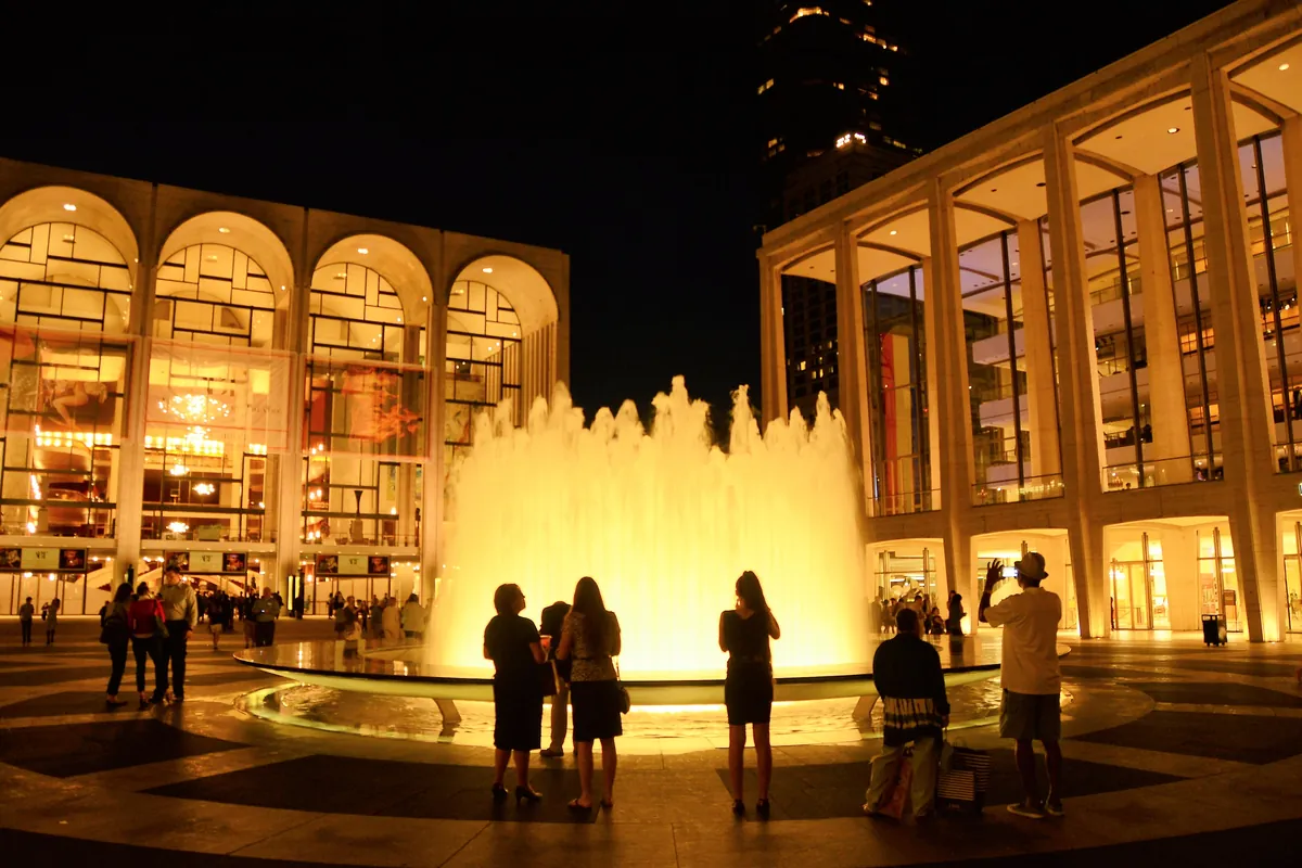 The Revson Fountain lit at night on the Lincoln Center plaza with the Metropolitan Opera House behind it