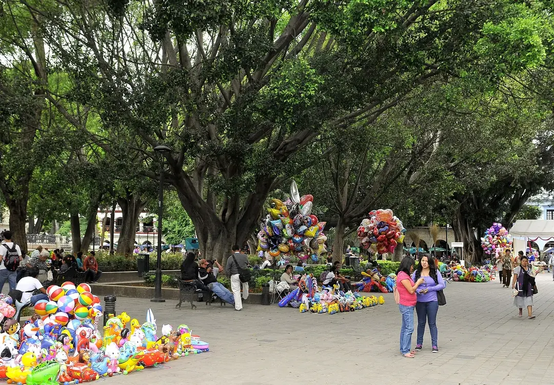 The Zócalo of Oaxaca de Juárez with crowds under tall laurel trees and a balloon seller