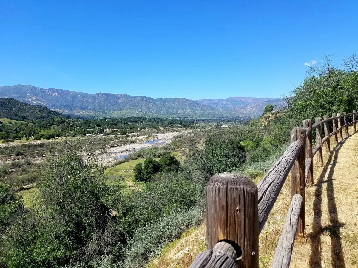A wooden fence along the Ventura River Parkway trail above the river valley, with mountains beyond