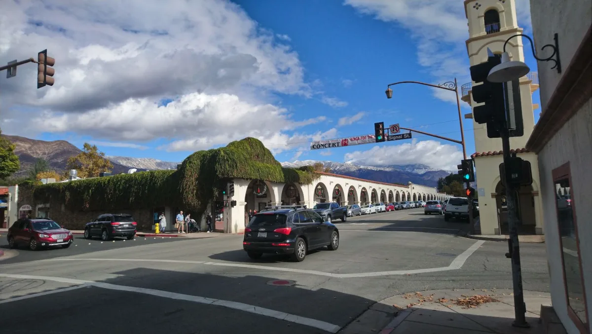 Ojai Avenue with the white arcade and its tower on the right, cars on the street and mountains under a blue sky