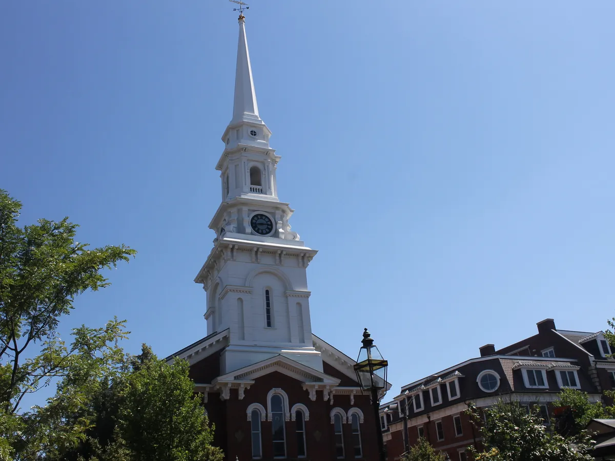The tall white steeple of the North Church against a blue sky above trees