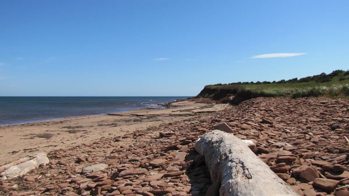 Cavendish Beach on Prince Edward Island, red sand and flat red stones below a low grassy bluff, with a driftwood log and blue sea