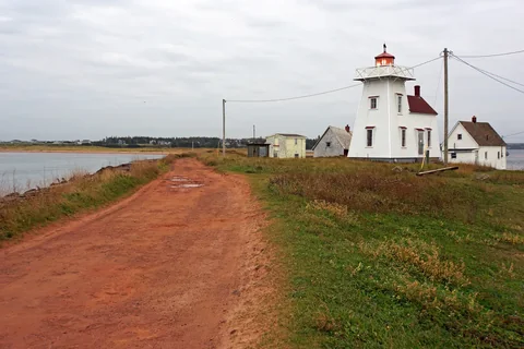 The North Rustico harbour lighthouse, a white tower attached to a house, at the end of a red dirt path