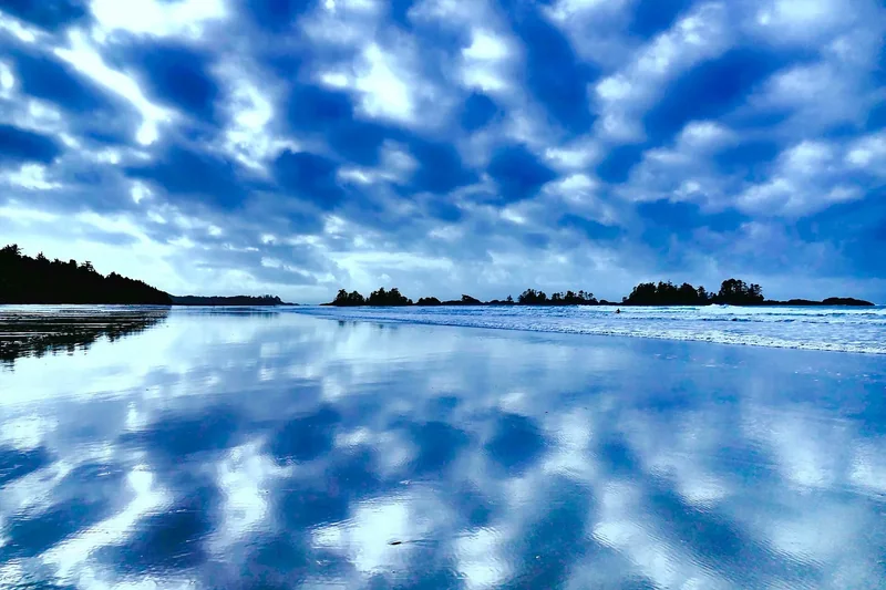 Clouds mirrored in wet sand on a wide Tofino beach, with dark forested islets on the horizon