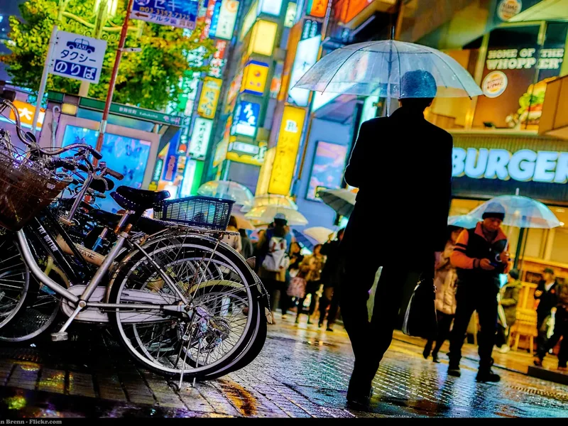 A rainy night street in Shinjuku with neon signs, bicycles and people holding umbrellas