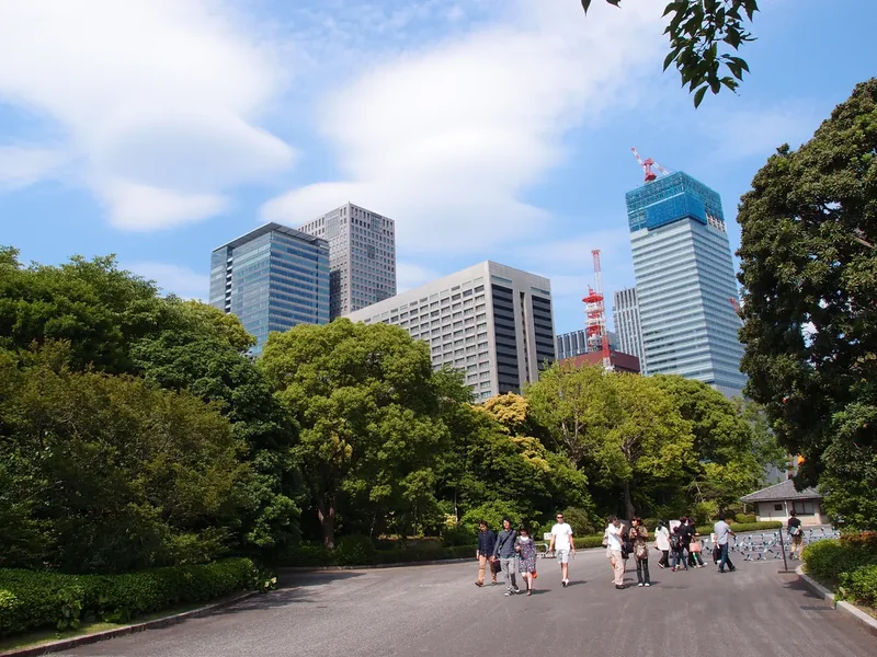 A wide gravel path through green trees in the Imperial Palace East Gardens, with office towers beyond