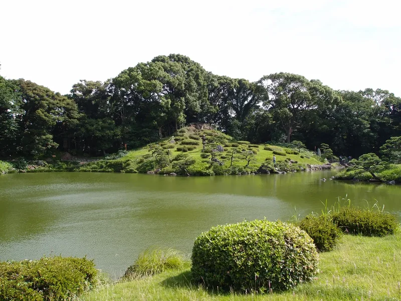 The pond at Kiyosumi Gardens with clipped shrubs, a grassy hill and trees reflected in the water